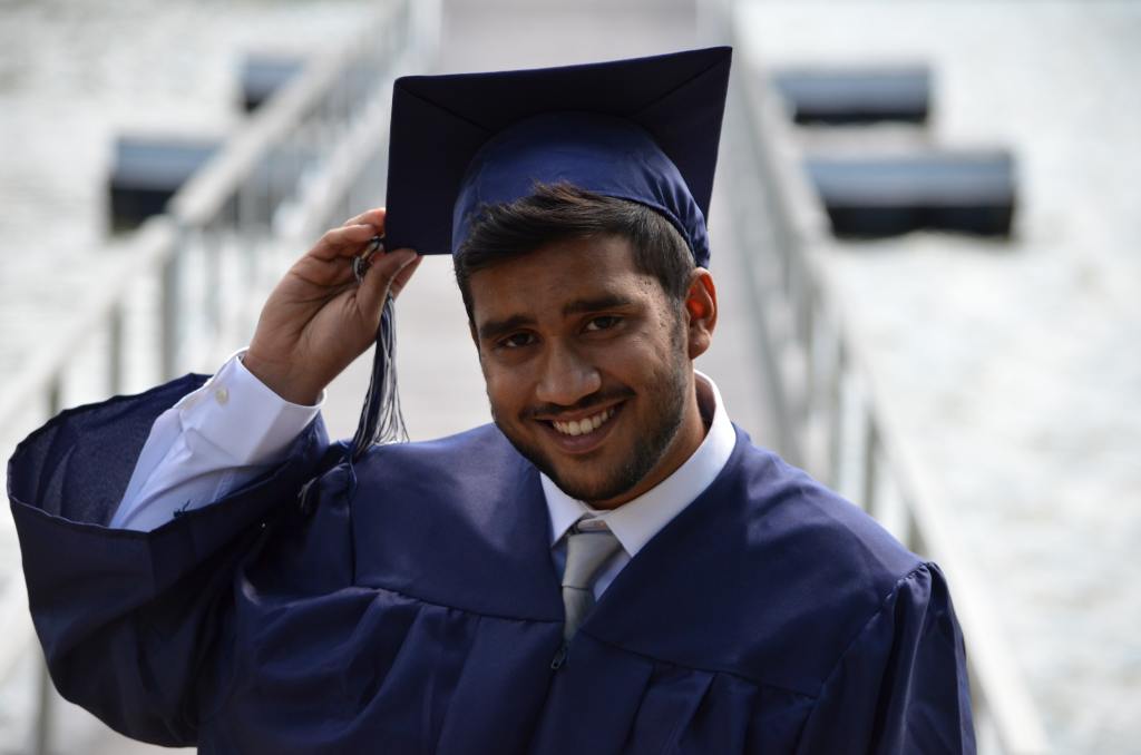 Decorative image: a person smiling at the camera wearing a mortarboard hat, which they are holding with their right hand.